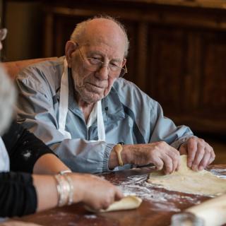 An elderly man wearing an apron, working with dough in a kitchen setting.