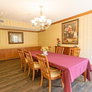 A classic dining room with a long table covered in a red tablecloth, surrounded by wooden chairs and illuminated by an elegant chandelier. Artwork hangs on the walls, enhancing the warm ambiance.