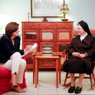 A conversation between a woman in casual attire and a nun in traditional religious dress, set in a cozy living room.