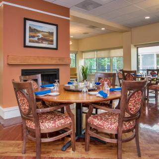 A cozy dining area with a well-set table and comfortable chairs, featuring warm interior decor and large windows.