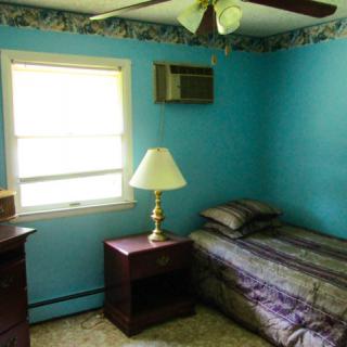 A small bedroom with blue walls featuring a single bed, a nightstand with a lamp, a window, and an air conditioner.