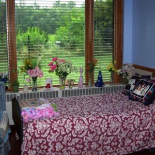 A cozy bedroom with a bed adorned with a floral cover, facing a large window with a view of lush greenery outside.