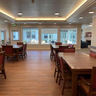 A spacious dining area with wooden tables and chairs, illuminated by ceiling lights and natural light from large windows.