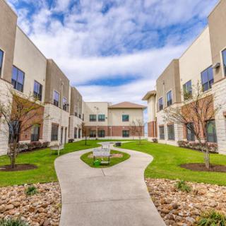 A serene courtyard surrounded by modern buildings, featuring a winding pathway and lush green grass under a partly cloudy sky.