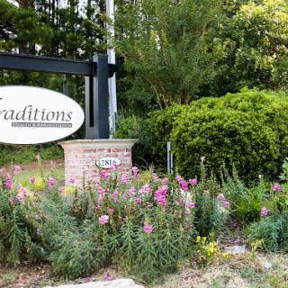 A welcoming sign for the community of Traditions, surrounded by vibrant flowers and lush greenery.