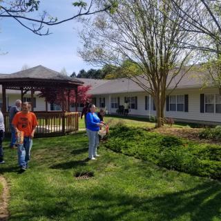 A group of people walking in a garden area with a gazebo, trees, and a building in the background on a sunny day.