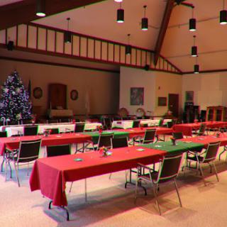 A spacious event room set up for a holiday gathering, featuring red and green tablecloths and a decorated Christmas tree.