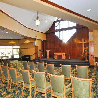 A chapel interior with rows of chairs, a podium on stage, and a large stained glass window.