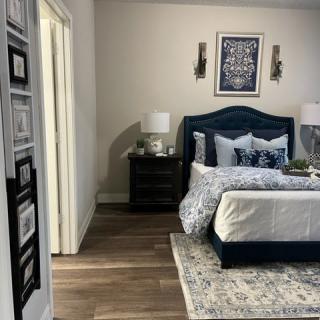 A well-decorated bedroom featuring a dark blue bed with patterned bedding, flanked by two bedside tables and lamps, complemented by artwork on the wall and a rug.