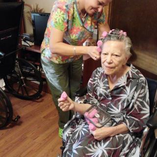 An elderly woman receiving hair styling assistance while seated in a wheelchair.