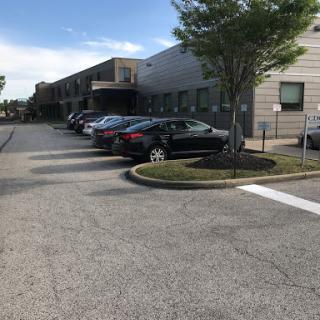 A view of a parking area with several cars parked alongside buildings and trees under a clear blue sky.