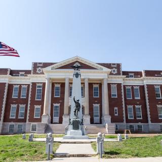 A historic building with columns and a statue in front, adorned by the American flag on a sunny day.