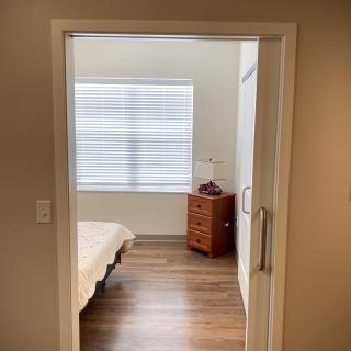 A minimalist bedroom with a bed and a wooden dresser, illuminated by natural light from a large window.