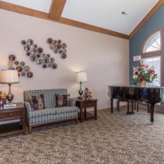 A cozy living room with a sofa, coffee tables, lamps, and a piano, featuring elegant wall art and carpeted flooring.