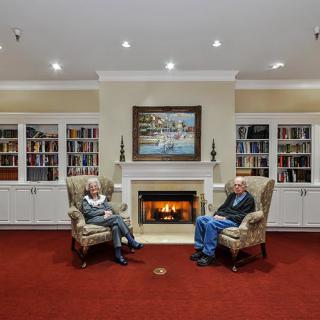 Two people sitting in armchairs by a fireplace in a library setting.