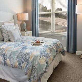 A cozy bedroom with a neatly made bed, featuring decorative pillows and a patterned comforter, is warmly lit by bedside lamps and natural light from the window.