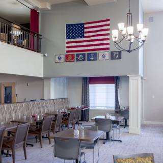 A spacious dining area featuring an American flag and military emblems on the wall, with a modern chandelier and neatly arranged tables and chairs.
