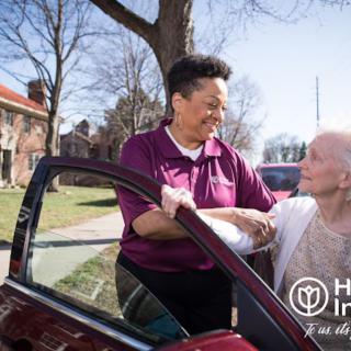 A caregiver assists an elderly woman getting out of a car, providing support and care outdoors.