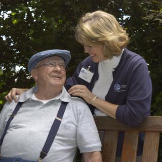 An elderly man sitting on a bench with a caregiver leaning over and interacting warmly outdoors.