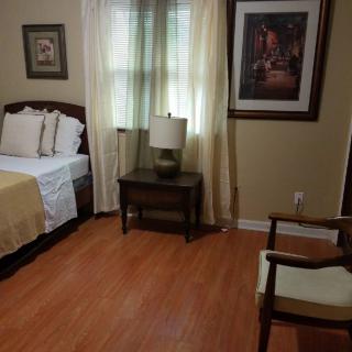 A cozy bedroom with a neatly made bed, side table with lamp, art on the walls and a wooden chair.
