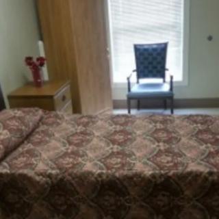 A simple bedroom with a patterned bedspread, chair by the window, and a nightstand with a vase.
