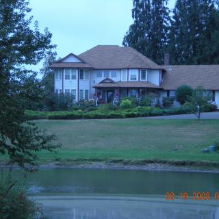 A large house with a sloped roof, surrounded by trees and greenery, overlooks a pond.