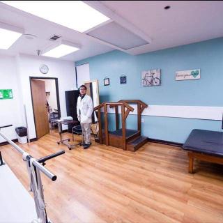 A physical therapy room equipped with exercise equipment, including parallel bars and a treadmill, with a healthcare professional present.