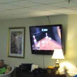 A living room scene featuring a wall-mounted television displaying a bowling video game, with nearby furniture and a lamp.