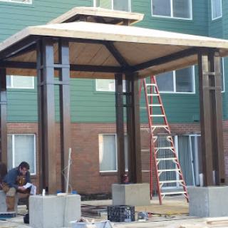 A partially constructed pavilion with a worker on-site, surrounded by scaffolding and building materials.