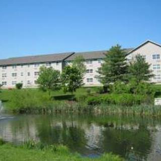 A large building sits beside a pond surrounded by trees and greenery under a clear blue sky.
