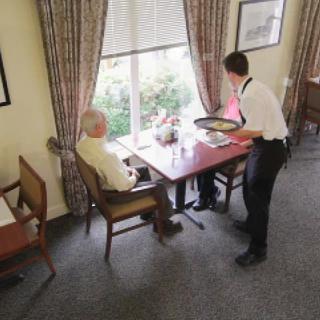 A waiter serves food to an elderly man seated at a table by the window in a restaurant.