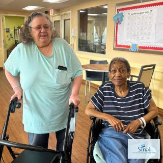 Two seniors are enjoying time together in a care facility, one seated in a wheelchair and the other using a walker.