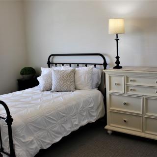 A minimalist bedroom with a metal-framed bed, white bedding, and a dresser topped with a lamp.