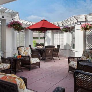 A cozy outdoor patio with comfortable seating and a red umbrella, surrounded by hanging flowers and a pergola.