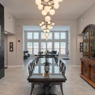 A spacious dining room featuring a long table with chairs, illuminated by modern chandeliers and natural light streaming in through large windows.
