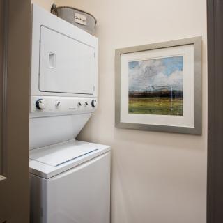 A small laundry area featuring a stacked washer and dryer unit with a framed landscape painting on the wall.