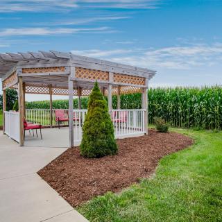 A charming gazebo in a rural setting surrounded by greenery and a cornfield under a blue sky.