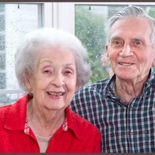 An elderly couple smiles warmly while sitting together by a window.