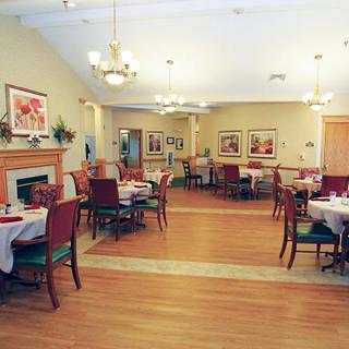 A cozy dining room with neatly arranged tables and chairs, featuring elegant chandeliers and a warm wooden floor.