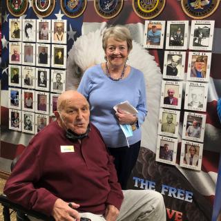 An elderly man in a wheelchair and a woman standing beside him, with a background displaying military portraits and symbols.