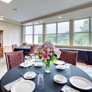 An elegantly set dining table with a beautiful floral centerpiece, surrounded by large windows that offer a scenic view.