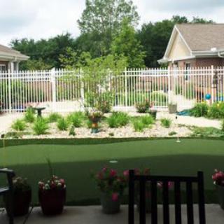 A well-maintained backyard garden with artificial grass, surrounded by a white fence and featuring various shrubs and plants.