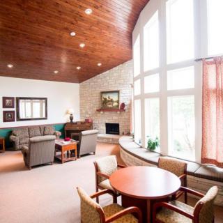 A well-lit living room featuring a cozy arrangement of sofas and chairs around a fireplace, with large windows and a wooden ceiling enhancing the inviting atmosphere.