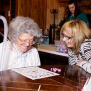 An elderly woman playing a game at a table with the assistance of another woman.