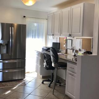 A modern kitchen with an integrated home office area, featuring white cabinets, a stainless steel refrigerator, and a desk with a computer.