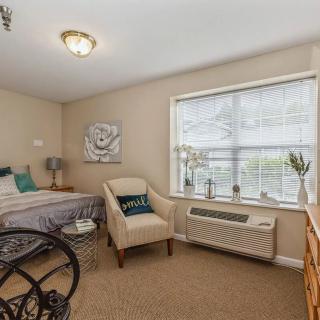 A cozy bedroom with a bed, an armchair, and a wheelchair by the window.