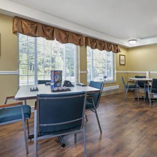 A bright dining room with tables and chairs arranged near large windows, featuring wooden flooring and decorative curtains.