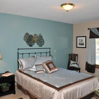 A cozy bedroom with a metal-frame bed, decorative pillows, and wall art above the headboard. A nightstand with a lamp sits beside the bed.