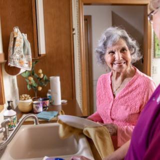 An elderly woman smiles while washing dishes in the kitchen.