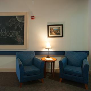 A cozy seating area with two blue armchairs, a small wooden table, and a lamp in between. A chalkboard and artwork decorate the wall.